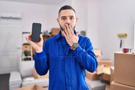 Photo for Hispanic man working on moving service showing smartphone screen covering mouth with hand, shocked and afraid for mistake. surprised expression - Royalty Free Image