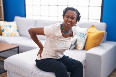 Téléchargez les photos : Moyen Age femme afro-américaine souffrant de maux de dos assis sur le canapé à la maison - en image libre de droit