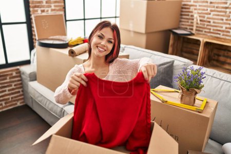 Foto de Mujer joven caucásica sonriendo confiado desembalaje ropa de cartón caja en nuevo hogar - Imagen libre de derechos