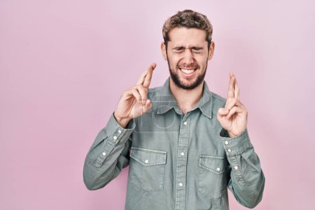Téléchargez les photos : Homme hispanique avec barbe debout sur fond rose doigt gestuel croisé souriant avec espoir et les yeux fermés. chance et concept superstitieux. - en image libre de droit