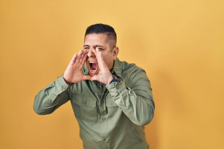 Téléchargez les photos : Jeune homme hispanique debout sur fond jaune criant en colère à haute voix avec les mains sur la bouche - en image libre de droit