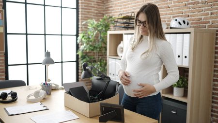 Photo for Young pregnant woman being fired packing belongings from workplace touching belly at office - Royalty Free Image