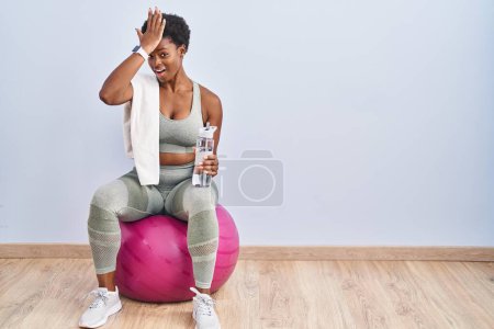 Téléchargez les photos : Femme afro-américaine portant des vêtements de sport assis sur pilates ballon surpris avec la main sur la tête pour erreur, rappelez-vous erreur. oublié, mauvais concept de mémoire. - en image libre de droit