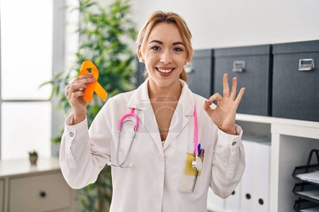 Téléchargez les photos : Médecin hispanique femme tenant conscience ruban orange faire ok signe avec les doigts, sourire geste amical excellent symbole - en image libre de droit