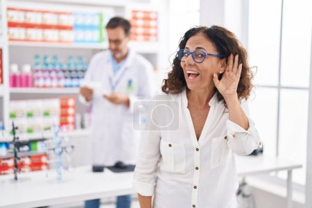 Foto de Mujer hispana de mediana edad comprando en farmacia sonriendo con la mano sobre el oído escuchando y escuchando rumores o chismes. concepto de sordera. - Imagen libre de derechos