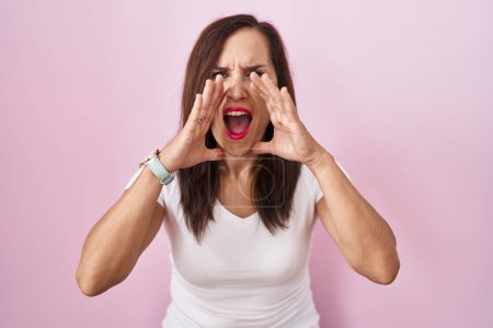 Téléchargez les photos : Moyen âge brunette femme debout sur fond rose criant en colère à haute voix avec les mains sur la bouche - en image libre de droit