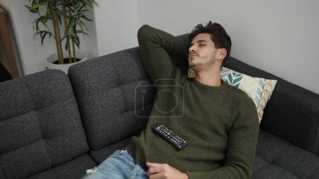 Photo for Young hispanic man holding tv remote control lying on sofa sleeping at home - Royalty Free Image