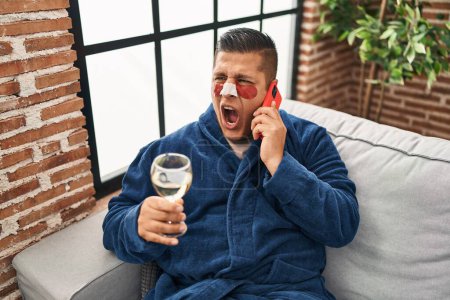 Photo for Hispanic young man wearing bathrobe and eye bags patches drinking wine speaking on the phone angry and mad screaming frustrated and furious, shouting with anger. rage and aggressive concept. - Royalty Free Image