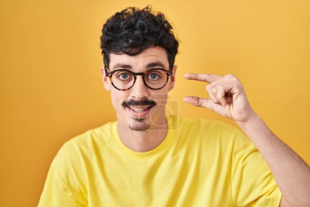 Téléchargez les photos : Homme hispanique portant des lunettes debout sur fond jaune geste souriant et confiant avec la main faisant signe de petite taille avec les doigts regardant et la caméra. concept de mesure. - en image libre de droit
