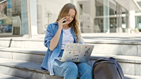 Foto de Joven estudiante rubia usando laptop hablando en smartphone en la universidad - Imagen libre de derechos