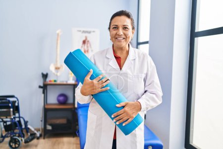 Foto de Middle age hispanic woman physiotherapist holding yoga mat at rehab clinic - Imagen libre de derechos