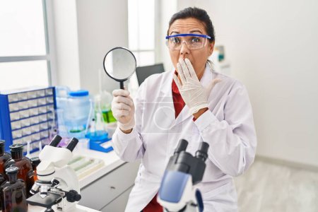 Photo for Middle age hispanic woman working at scientist laboratory holding magnifying glass covering mouth with hand, shocked and afraid for mistake. surprised expression - Royalty Free Image