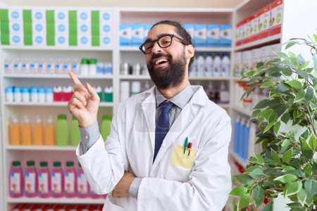 Photo for Hispanic man with beard working at pharmacy drugstore smiling happy pointing with hand and finger to the side - Royalty Free Image