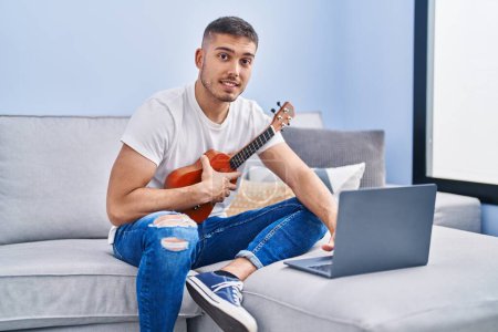 Photo for Young hispanic man having online ukelele class sitting on sofa at home - Royalty Free Image
