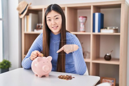 Photo for Chinese young woman putting coin in piggy bank smiling happy pointing with hand and finger - Royalty Free Image