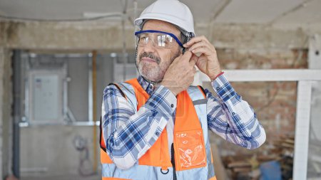 Foto de Constructor hombre de mediana edad sonriendo confiado usando hardhat en el sitio de construcción - Imagen libre de derechos