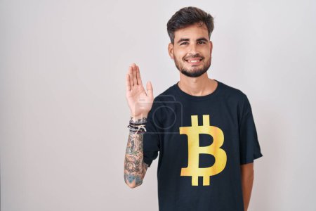 Téléchargez les photos : Jeune homme hispanique avec des tatouages portant Bitcoin t-shirt renonçant à dire bonjour heureux et souriant, geste de bienvenue convivial - en image libre de droit