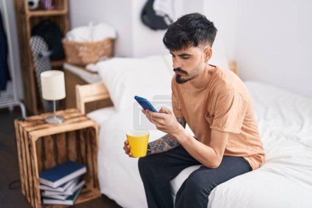 Photo for Young hispanic man using smartphone drinking coffee at bedroom - Royalty Free Image