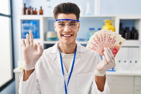 Photo for Young hispanic man working at scientist laboratory holding shekels doing ok sign with fingers, smiling friendly gesturing excellent symbol - Royalty Free Image