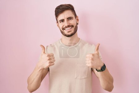 Hispanic man with beard standing over pink background success sign doing positive gesture with hand, thumbs up smiling and happy. cheerful expression and winner gesture.