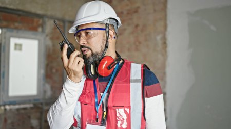 Téléchargez les photos : Jeune homme latin constructeur parler sur walkie-talkie sur le chantier de construction - en image libre de droit