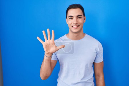 Téléchargez les photos : Jeune homme hispanique debout sur fond bleu montrant et pointant vers le haut avec les doigts numéro cinq tout en souriant confiant et heureux. - en image libre de droit