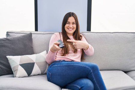 Téléchargez les photos : Jeune femme brune tenant la télévision télécommande souriant heureux pointant avec la main et le doigt - en image libre de droit
