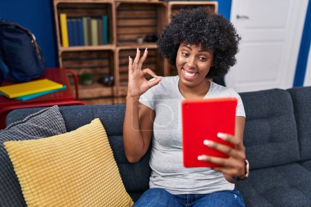 Téléchargez les photos : Femme noire avec les cheveux bouclés en utilisant le pavé tactile assis sur le canapé faisant signe ok avec les doigts, sourire geste amical excellent symbole - en image libre de droit