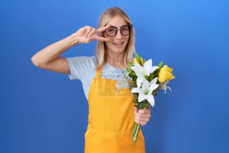 Téléchargez les photos : Jeune femme caucasienne portant tablier de fleuriste tenant des fleurs faisant symbole de paix avec les doigts sur le visage, souriant joyeuse montrant la victoire - en image libre de droit