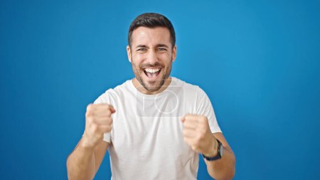 Téléchargez les photos : Jeune homme hispanique souriant confiant avec expression gagnante sur fond bleu isolé - en image libre de droit