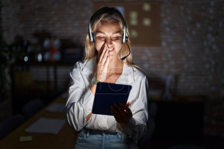Photo for Young blonde woman working at the office at night laughing and embarrassed giggle covering mouth with hands, gossip and scandal concept - Royalty Free Image