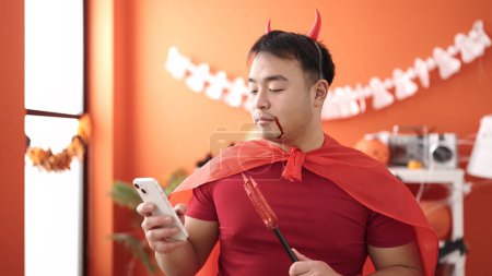 Téléchargez les photos : Jeune homme chinois portant costume diable à l'aide d'un smartphone à la maison - en image libre de droit