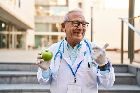Téléchargez les photos : Médecin sénior aux cheveux gris tenant la pomme verte saine pointant le pouce vers le côté souriant heureux avec la bouche ouverte - en image libre de droit