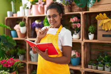 Téléchargez les photos : Afro-américaine femme fleuriste souriant confiant carnet de lecture à la boutique de fleurs - en image libre de droit