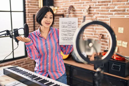 Téléchargez les photos : Jeune femme asiatique faisant tutoriel de musique en ligne montrant feuille de musique célébrant la réalisation avec sourire heureux et expression gagnante avec la main levée - en image libre de droit