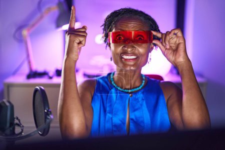 African woman with dreadlocks playing video games wearing virtual reality glasses smiling with an idea or question pointing finger with happy face, number one