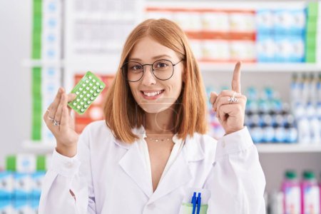 Photo for Young redhead woman working at pharmacy drugstore holding birth control pills surprised with an idea or question pointing finger with happy face, number one - Royalty Free Image