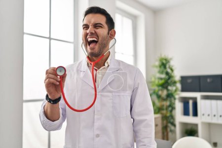 Photo for Young hispanic doctor man with beard holding stethoscope auscultating angry and mad screaming frustrated and furious, shouting with anger. rage and aggressive concept. - Royalty Free Image