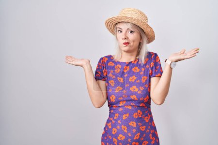 Photo for Young caucasian woman wearing flowers dress and summer hat clueless and confused expression with arms and hands raised. doubt concept. - Royalty Free Image