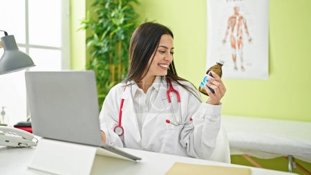 Téléchargez les photos : Jeune belle hispanique femme médecin en utilisant ordinateur portable tenant bouteille de médicaments à la clinique - en image libre de droit