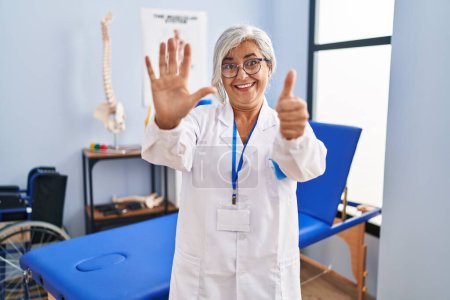 Foto de Mujer de mediana edad con el pelo gris trabajando en la clínica de recuperación del dolor mostrando y apuntando hacia arriba con los dedos número seis mientras sonríe confiado y feliz. - Imagen libre de derechos