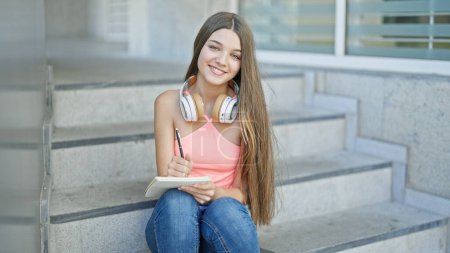 Téléchargez les photos : Jeune belle étudiante prenant des notes assis sur les escaliers souriant à l'école - en image libre de droit