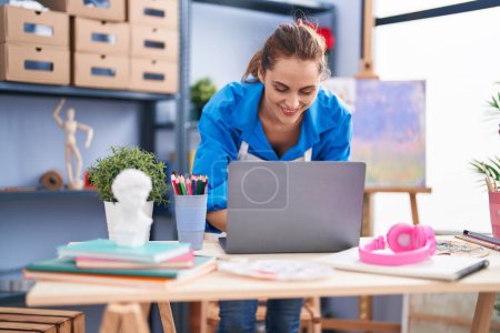 Téléchargez les photos : Jeune femme artiste souriant confiant en utilisant un ordinateur portable au studio d'art - en image libre de droit