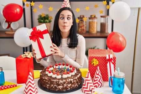 Foto de Mujer hispana celebrando cumpleaños con pastel sosteniendo regalo haciendo cara de pez con boca y ojos entrecerrados, loco y cómico. - Imagen libre de derechos