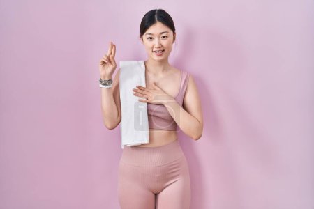 Téléchargez les photos : Jeune femme chinoise portant des vêtements de sport et une serviette souriante jurant avec la main sur la poitrine et les doigts vers le haut, faisant un serment de fidélité - en image libre de droit