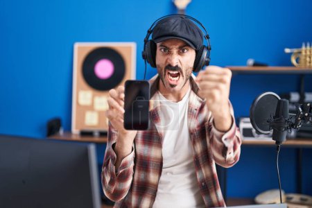 Téléchargez les photos : Homme hispanique avec barbe au studio de musique montrant smartphone ennuyé et frustré criant de colère, criant fou de colère et la main levée - en image libre de droit
