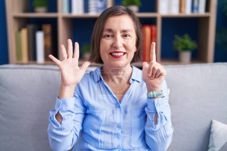 Téléchargez les photos : Moyen Age femme hispanique assis sur le canapé à la maison montrant et pointant vers le haut avec les doigts numéro sept tout en souriant confiant et heureux. - en image libre de droit