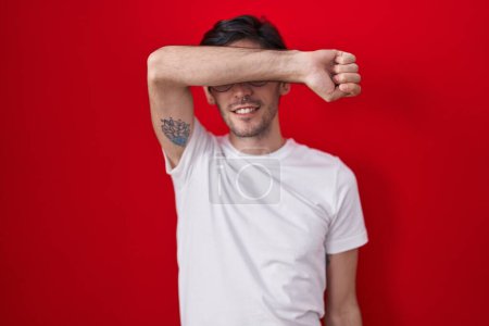 Téléchargez les photos : Jeune homme hispanique debout sur fond rouge couvrant les yeux avec le bras souriant gai et drôle. concept aveugle. - en image libre de droit