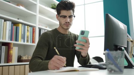 Téléchargez les photos : Young hispanic man student using smartphone writing on notebook at library university - en image libre de droit