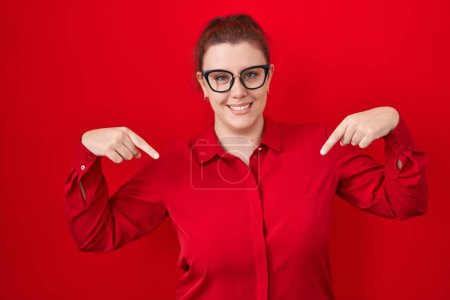 Foto de Mujer hispana joven con el pelo rojo de pie sobre fondo rojo mirando confiado con sonrisa en la cara, señalándose con los dedos orgullosos y felices. - Imagen libre de derechos